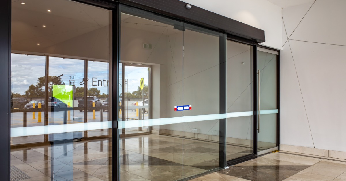 A modern glass sliding door entrance with metal side panels, reflective windows, and smooth stone flooring.
