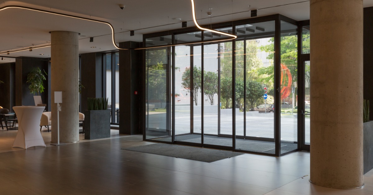 Inside a hotel lobby, there is a glass automatic sliding door entrance with metal panels and tile flooring.