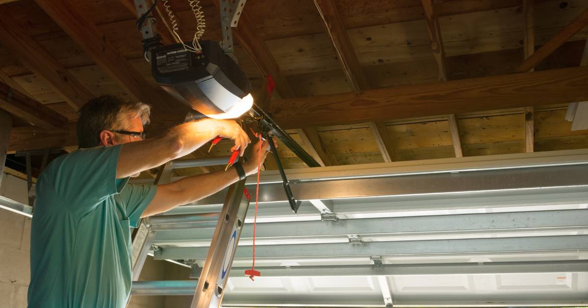 A man wearing safety glasses while standing on a ladder and using a tool to work on a residential garage door.