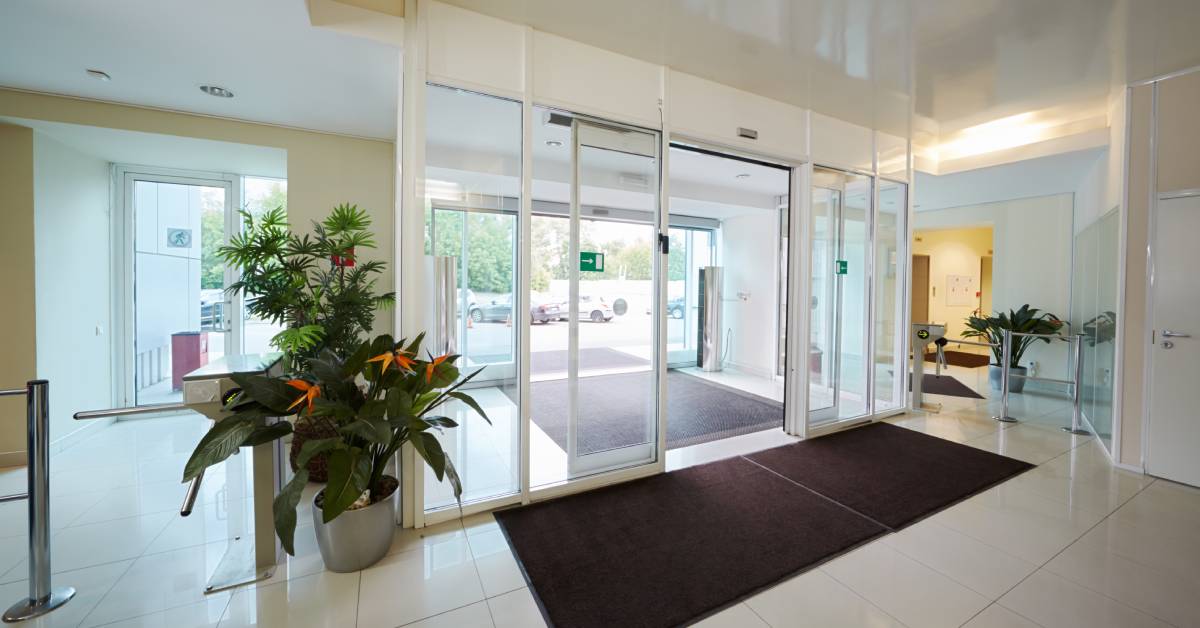 A building entrance equipped with glass automatic doors. There are large potted plants next to the doors.