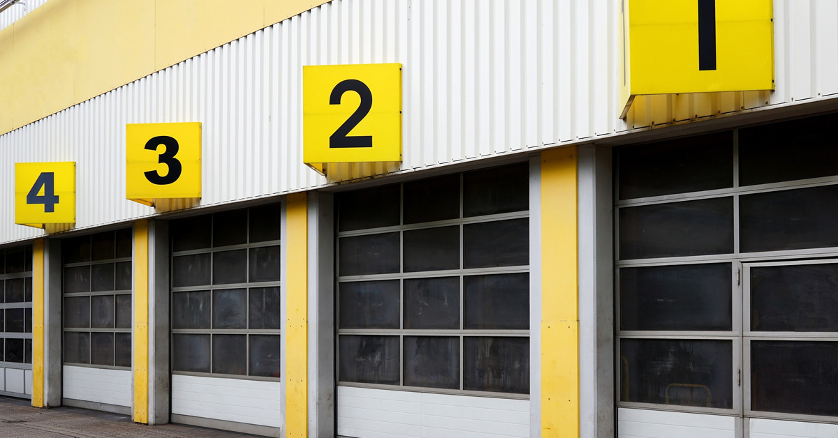 The exterior of a warehouse-style building with several roll-up doors in a row. The doors are numbered with yellow signs.