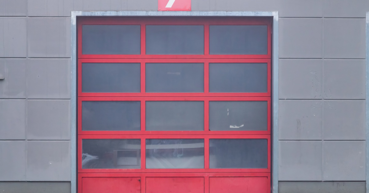 A red garage door with several small windows and a sign above it with the number seven attached to a gray building.