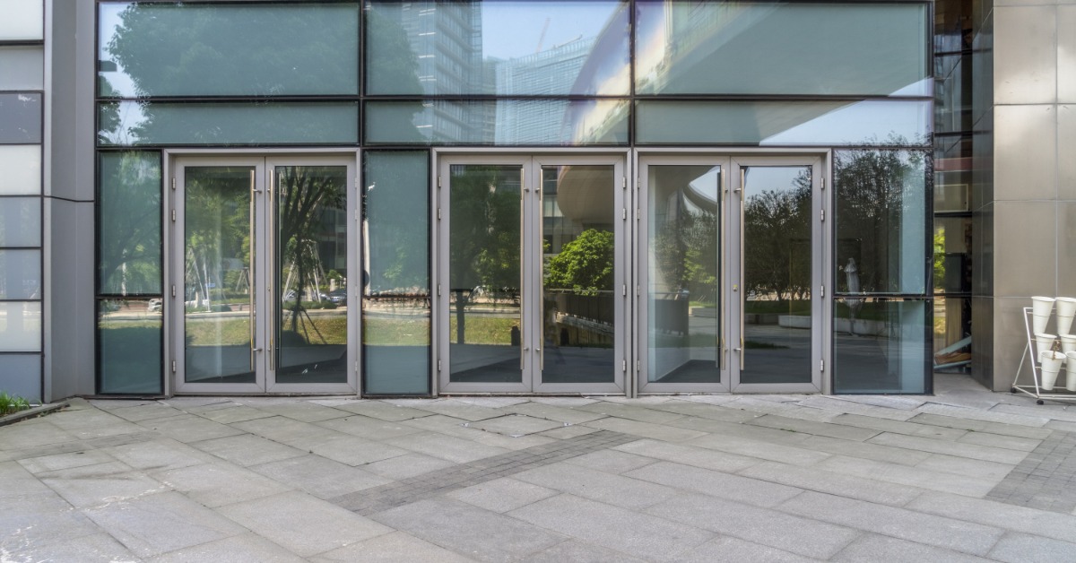 Three sets of glass commercial doors outside of a building. There is a concrete walkway in front of the doors.