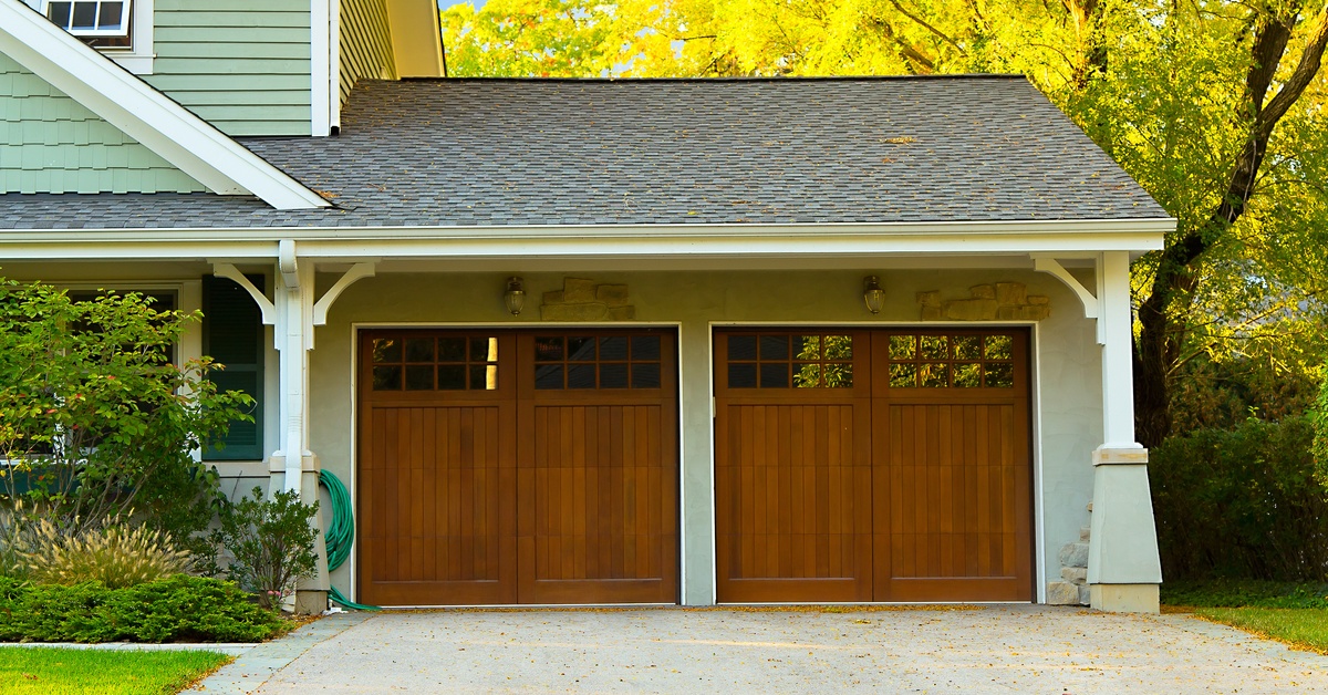 Two wooden garage doors that are connected to a house with green siding. There are windows built into the garage doors.