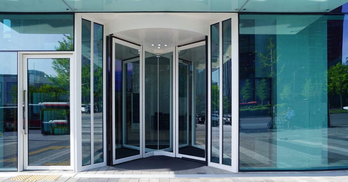 A glass revolving door at a modern building entrance with blue-tinted windows, white columns, and a paved walkway.