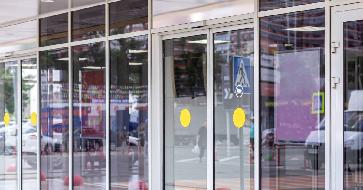 Clear glass doors lead into a supermarket. The doors have no handles but have a sensor to slide open upon entry.