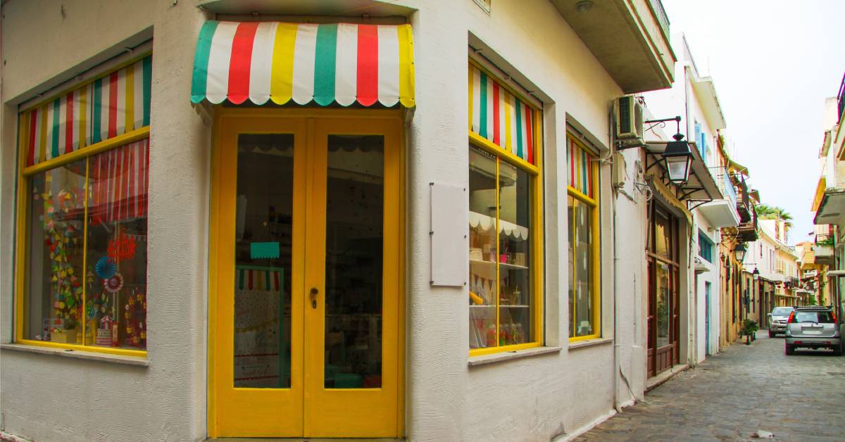 A small yellow door on the corner of a street. Above the door is a multicolored awning with matching window shades.